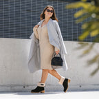 Žena v šatách a kabáte nosí vysoké izolované topánky NOEL, kráča po meste s kabelkou.