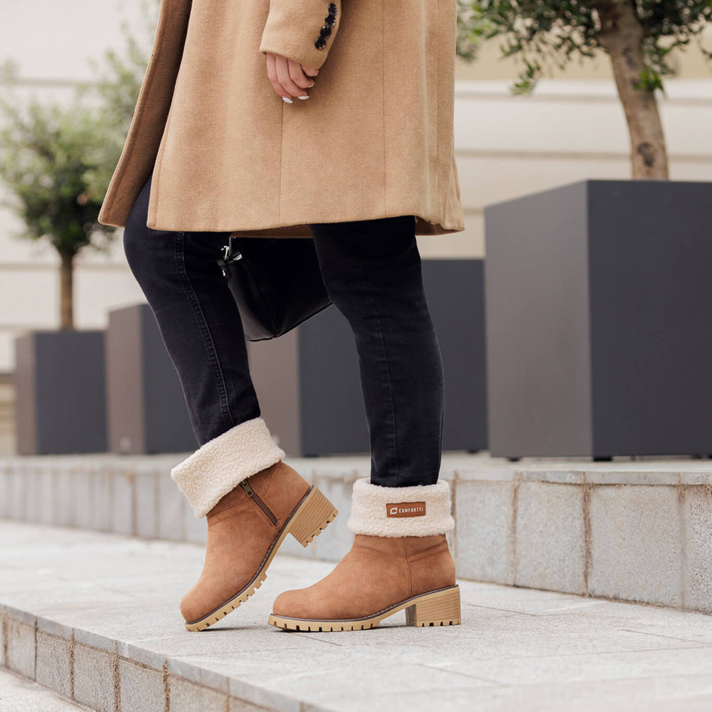 Osoba v kabáte obúva vysoké izolované topánky NOEL v hnedej farbe na mestskom chodníku.